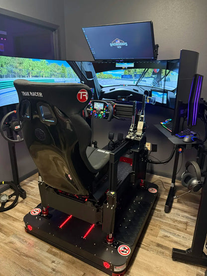 A high-end truck simulator racing shell with a black bucket seat, steering wheel, pedals, and multiple monitors showing a virtual race track. The setup is placed in a room with wood flooring and dark gray walls.